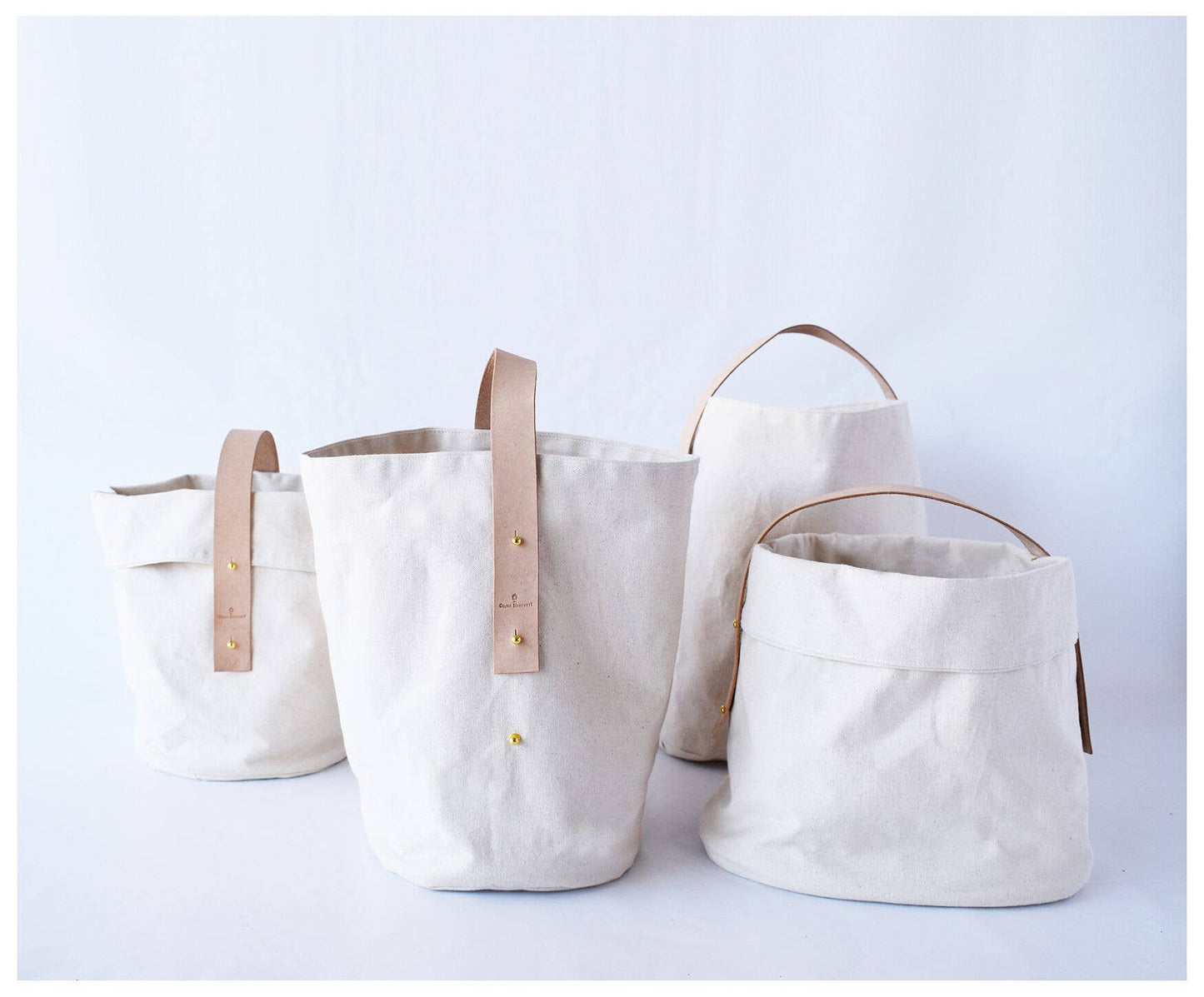 4 canvas baskets side by side. Two of the canvas baskets are folded down at the top to shorten. All baskets have a leather strap with brass buttons.